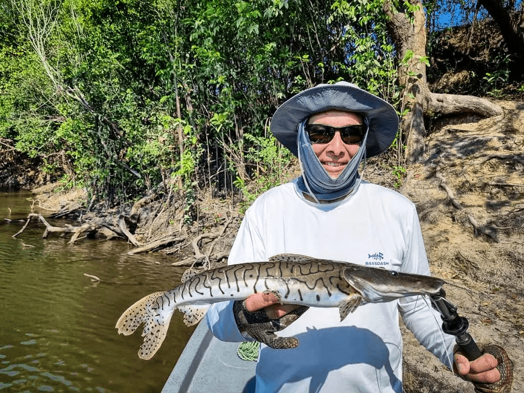 Pescador con sombrero sostiene un bagre rayado de manchas negras sobre la lancha.