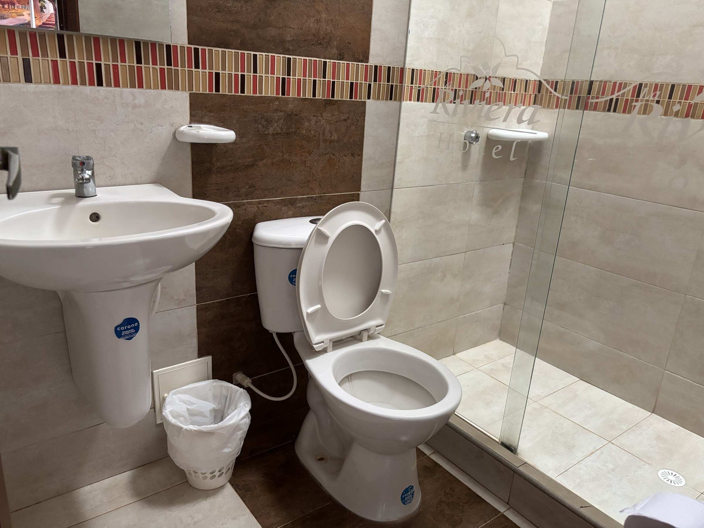 Baño con lavamanos, sanitario y ducha de vidrio, con una franja de enchape de colores.