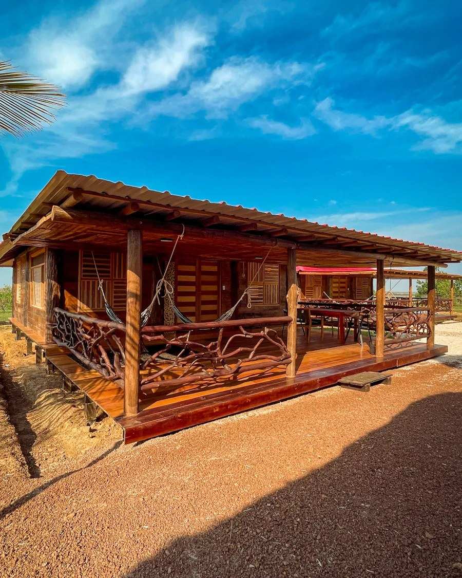 Cabaña de madera con corredor de barandas de troncos y hamacas, bajo el cielo azul.