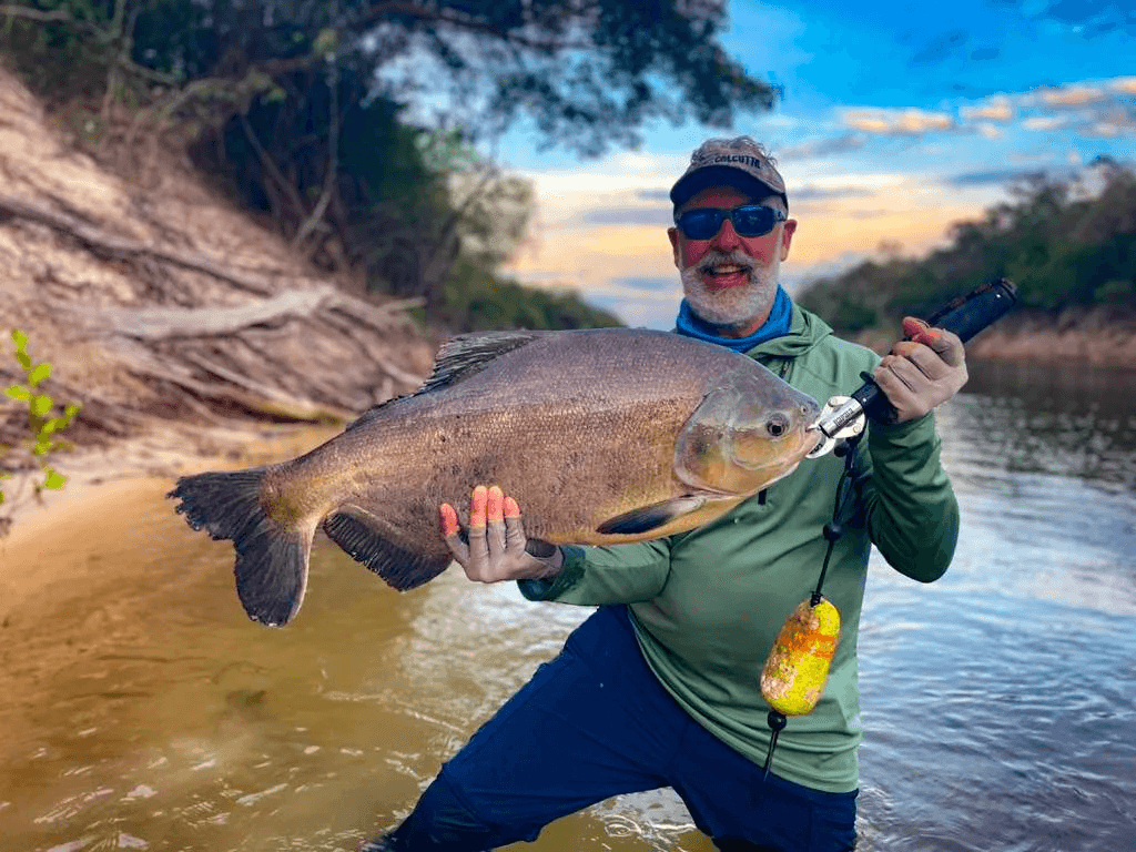 Pescador de gafas sostiene una cachama oscura y redonda junto al río.