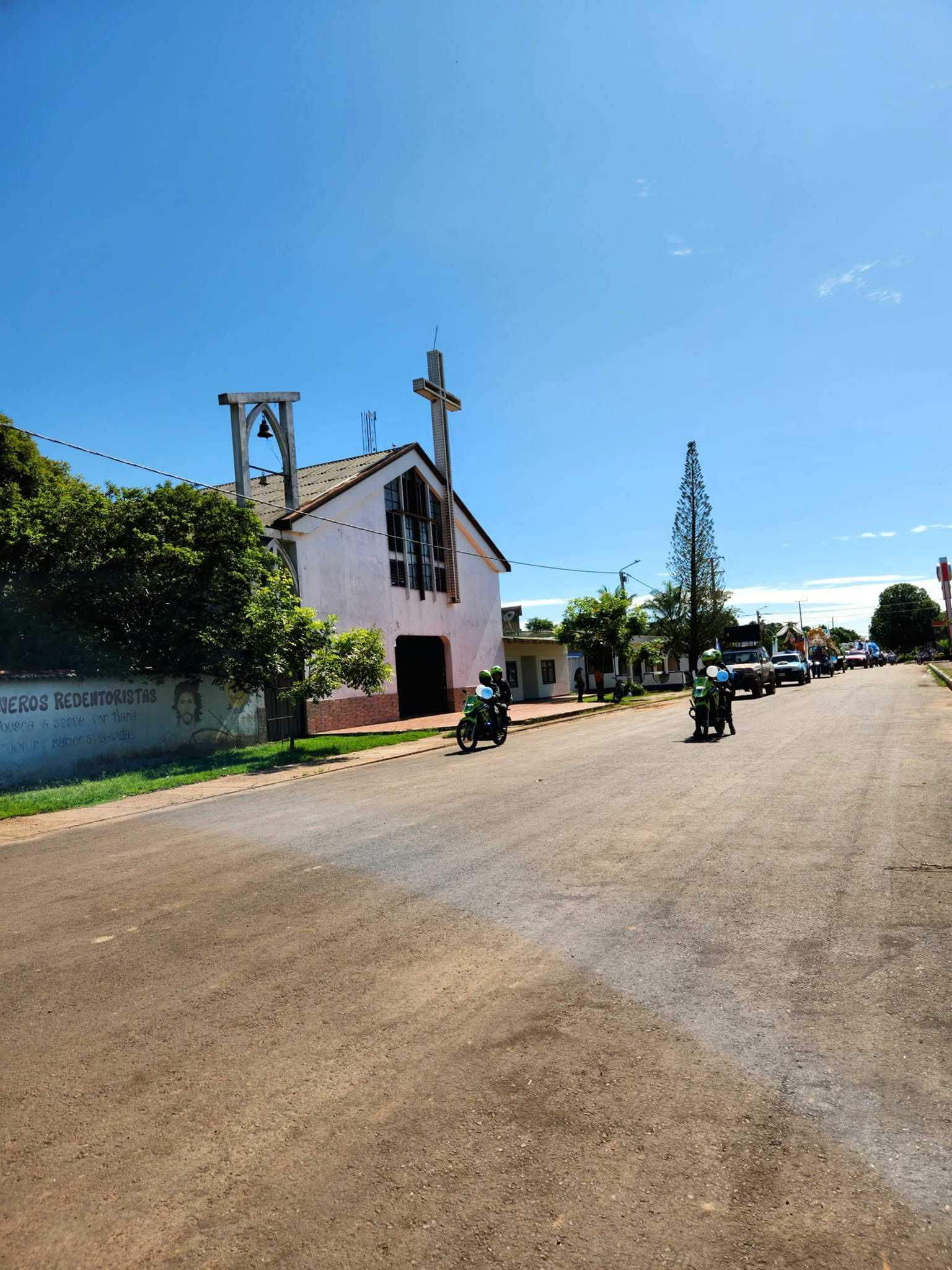 La iglesia blanca de La Primavera junto a la calle por donde pasa un desfile de motos y camionetas.