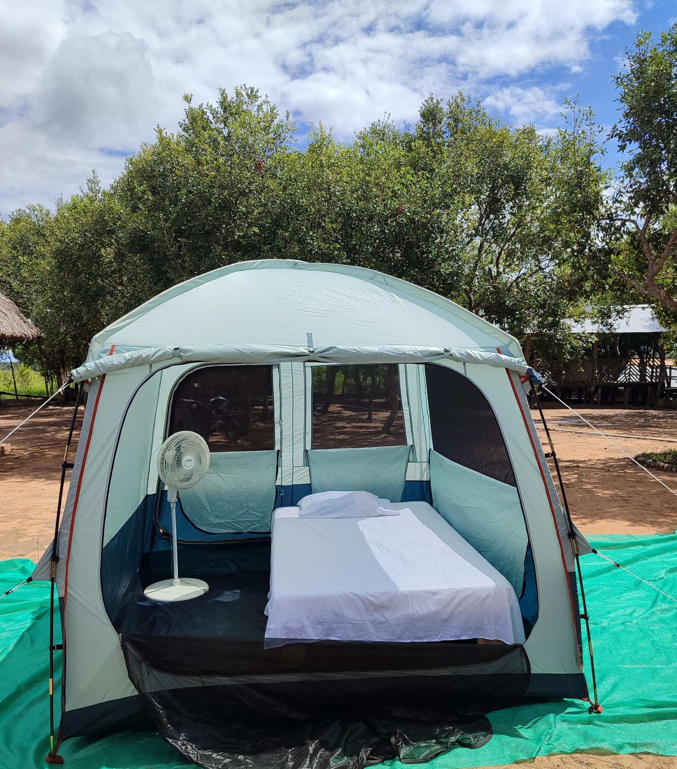 Carpa amplia con cama tendida y ventilador, sobre una lona verde.
