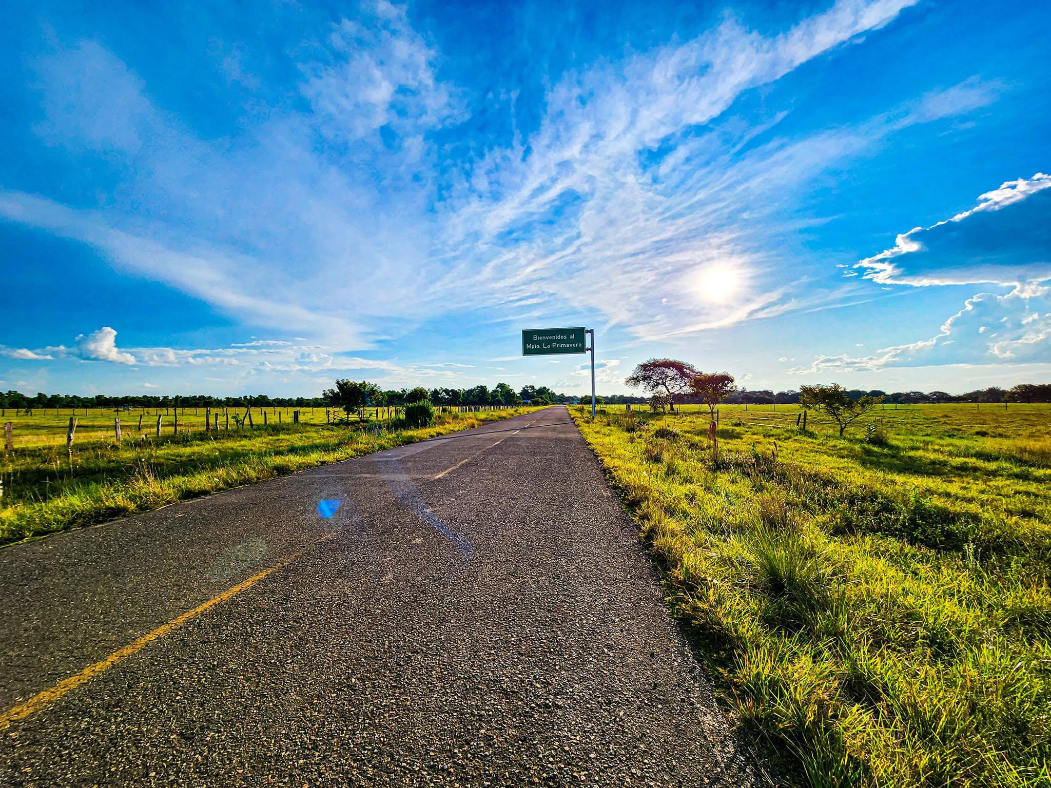 La carretera recta entre la sabana verde, bajo un cielo azul, con el letrero de bienvenida a lo lejos.