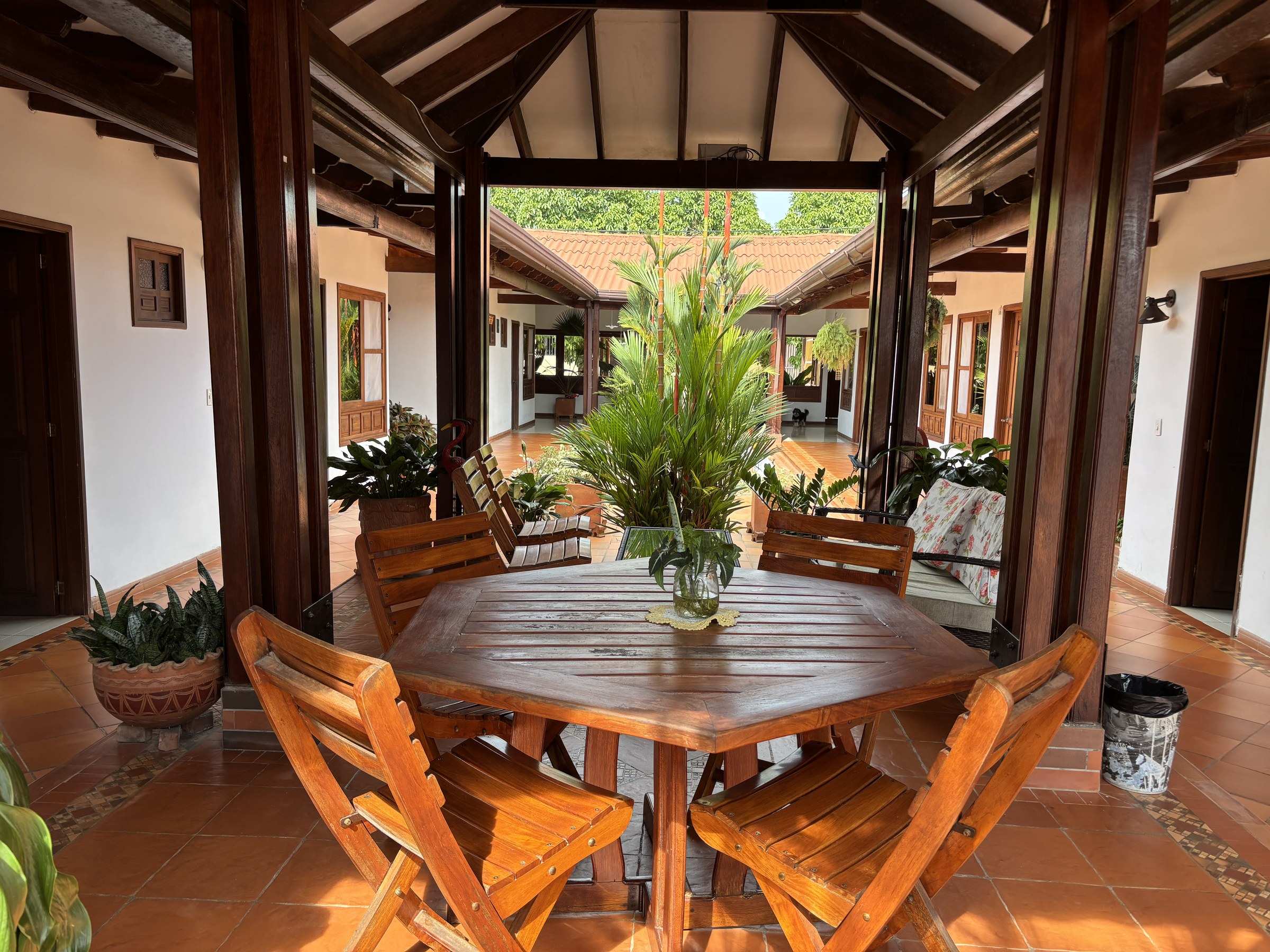 Mesa de madera con sillas plegables bajo una cubierta de vigas, en medio del corredor que da al patio.