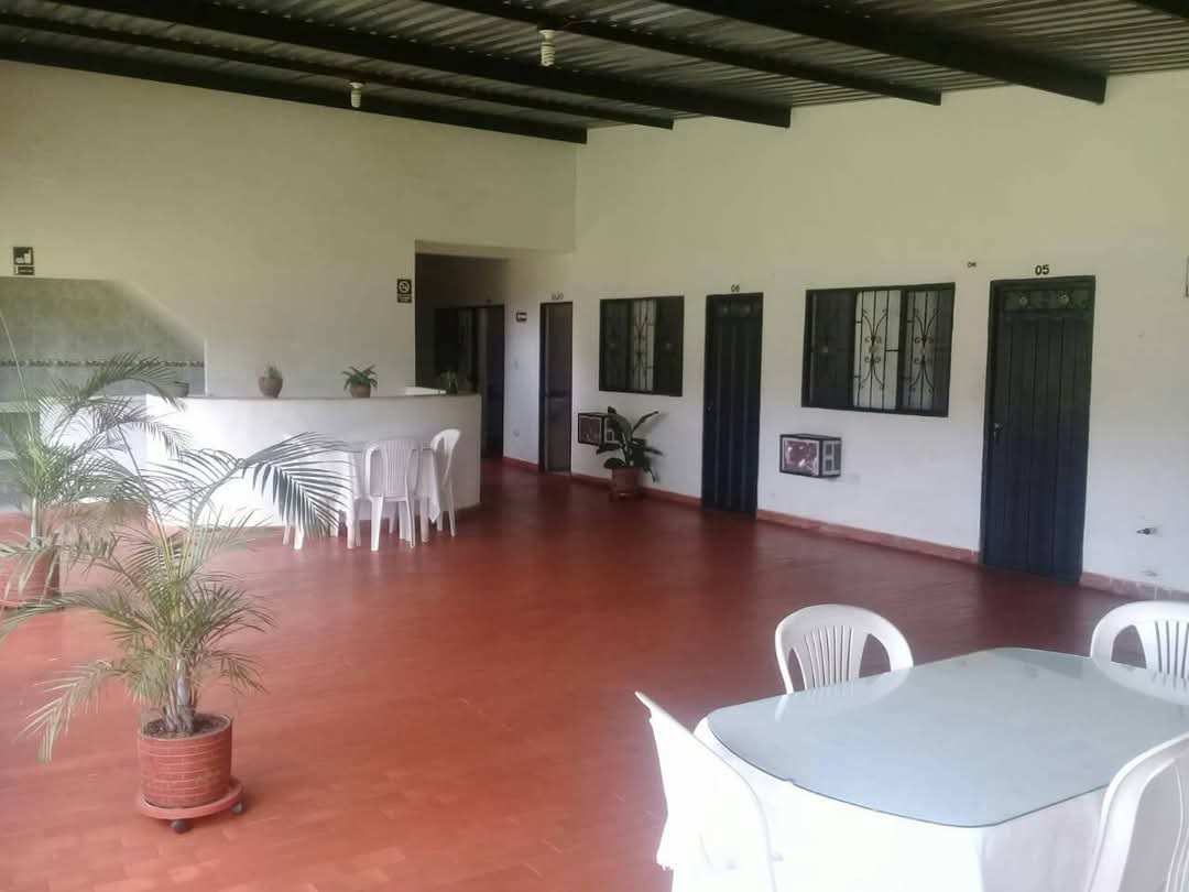 Corredor interior de piso rojo con puertas oscuras a las habitaciones, mesas y sillas blancas, palmas en materas y un mostrador blanco al fondo.