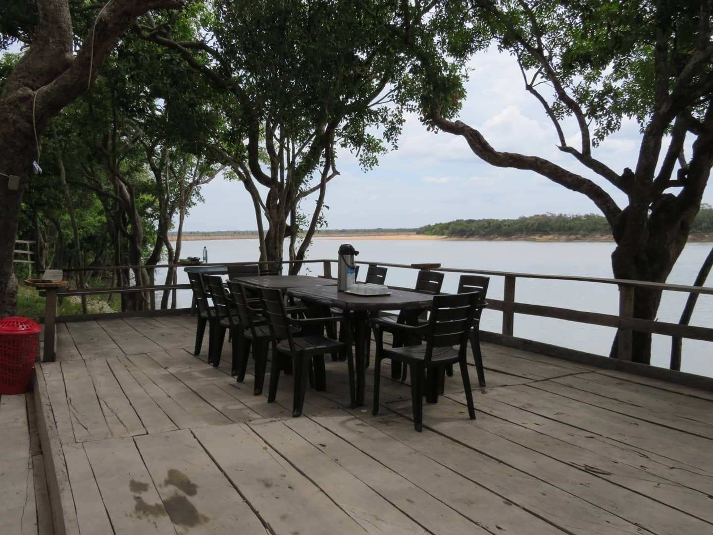 Deck de madera con mesas y sillas bajo los árboles, frente al río Tomo.
