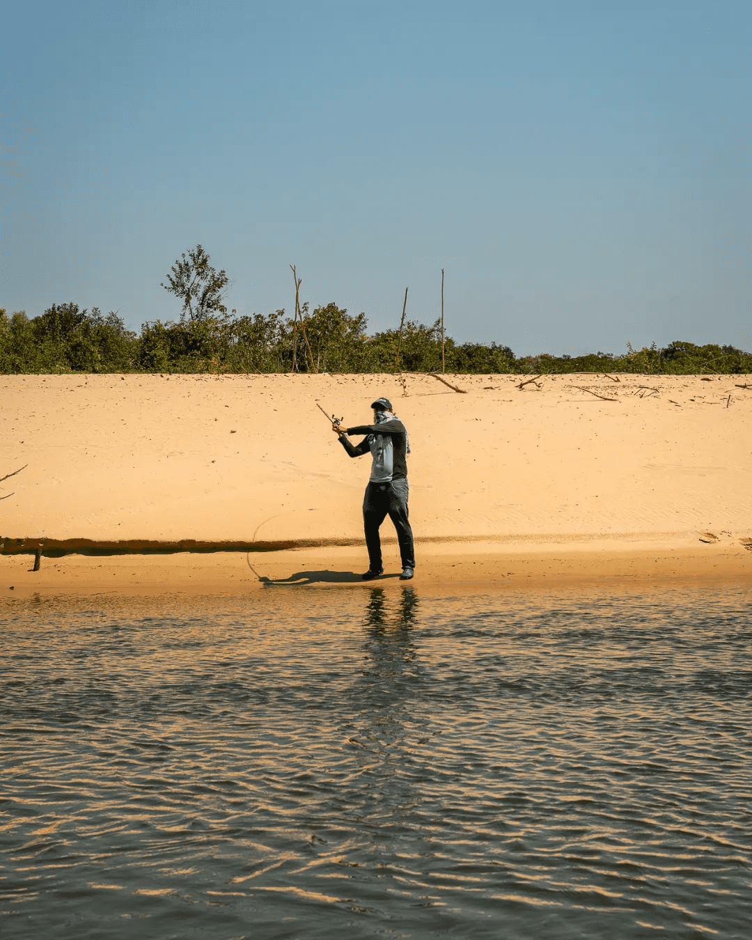 Pescador lanza la línea de pie en una playa de arena del río, con el monte detrás.