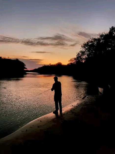 Silueta de un pescador que lanza desde la orilla del río bajo un cielo de atardecer.