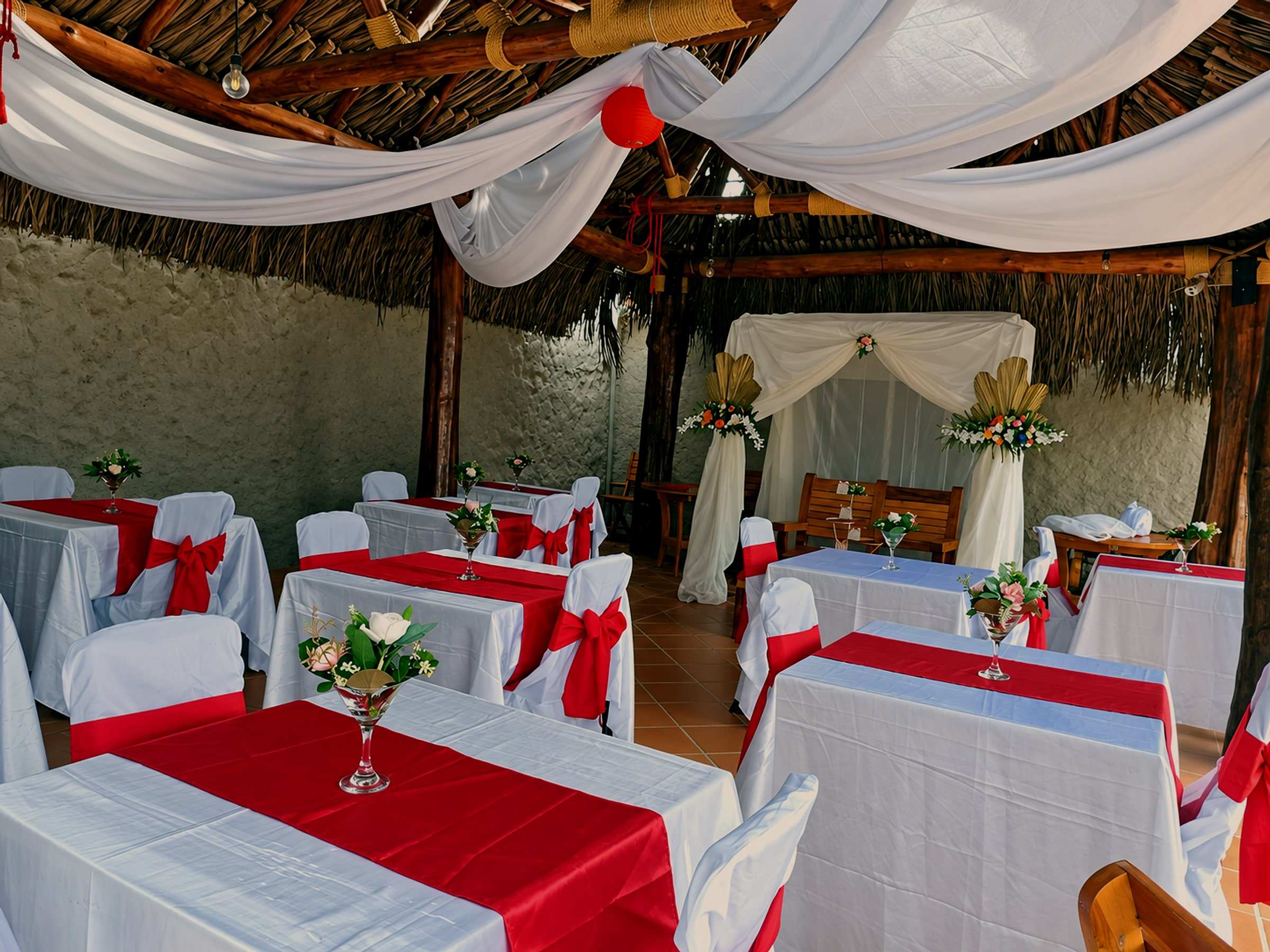 Mesas con manteles blancos y rojos bajo telas blancas, con un arco de flores al fondo.