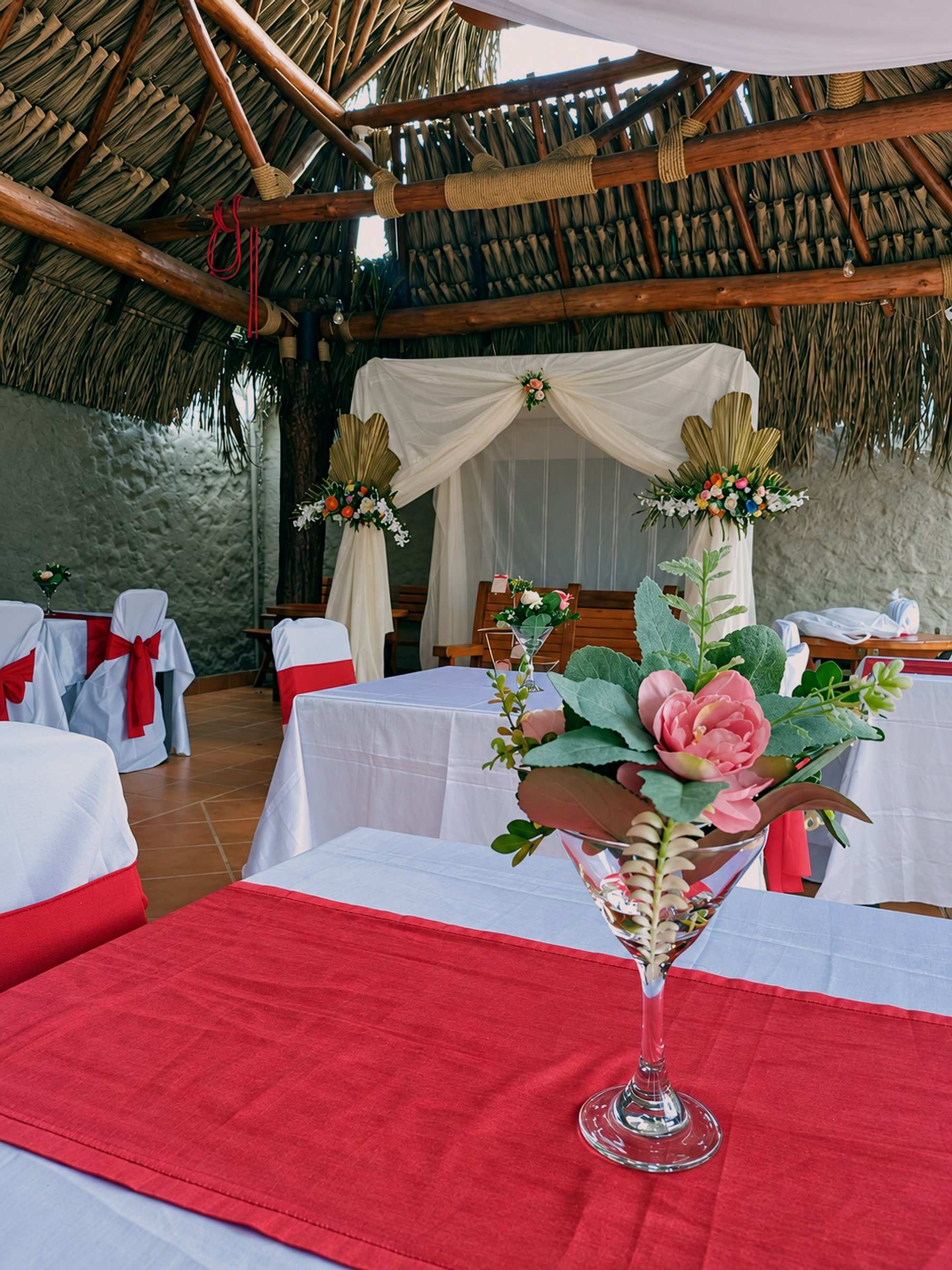 Centro de mesa con rosas en una copa sobre un camino rojo, con el salón decorado detrás.