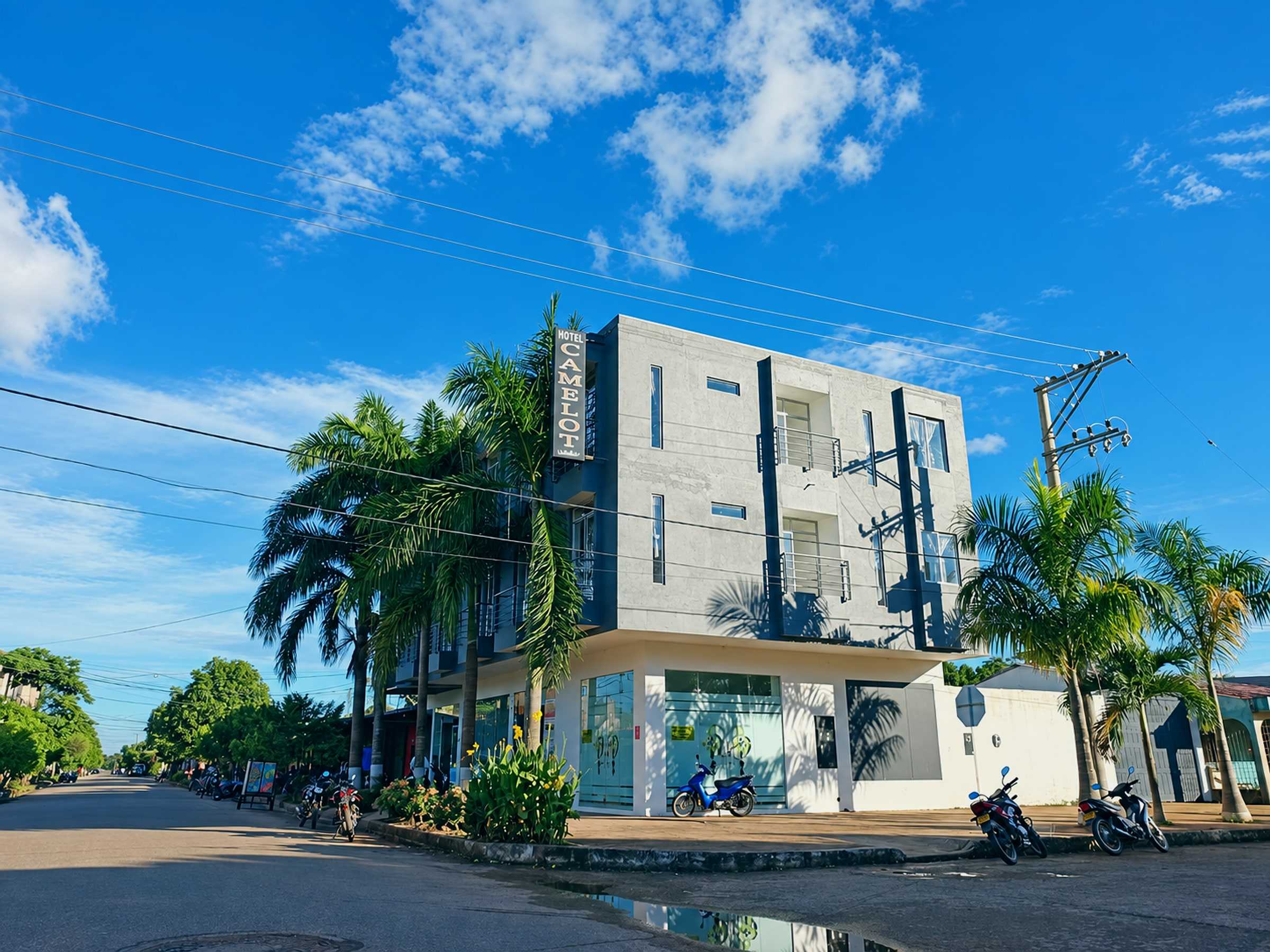 Esquina del hotel bajo un cielo azul, con palmas y motos al frente.