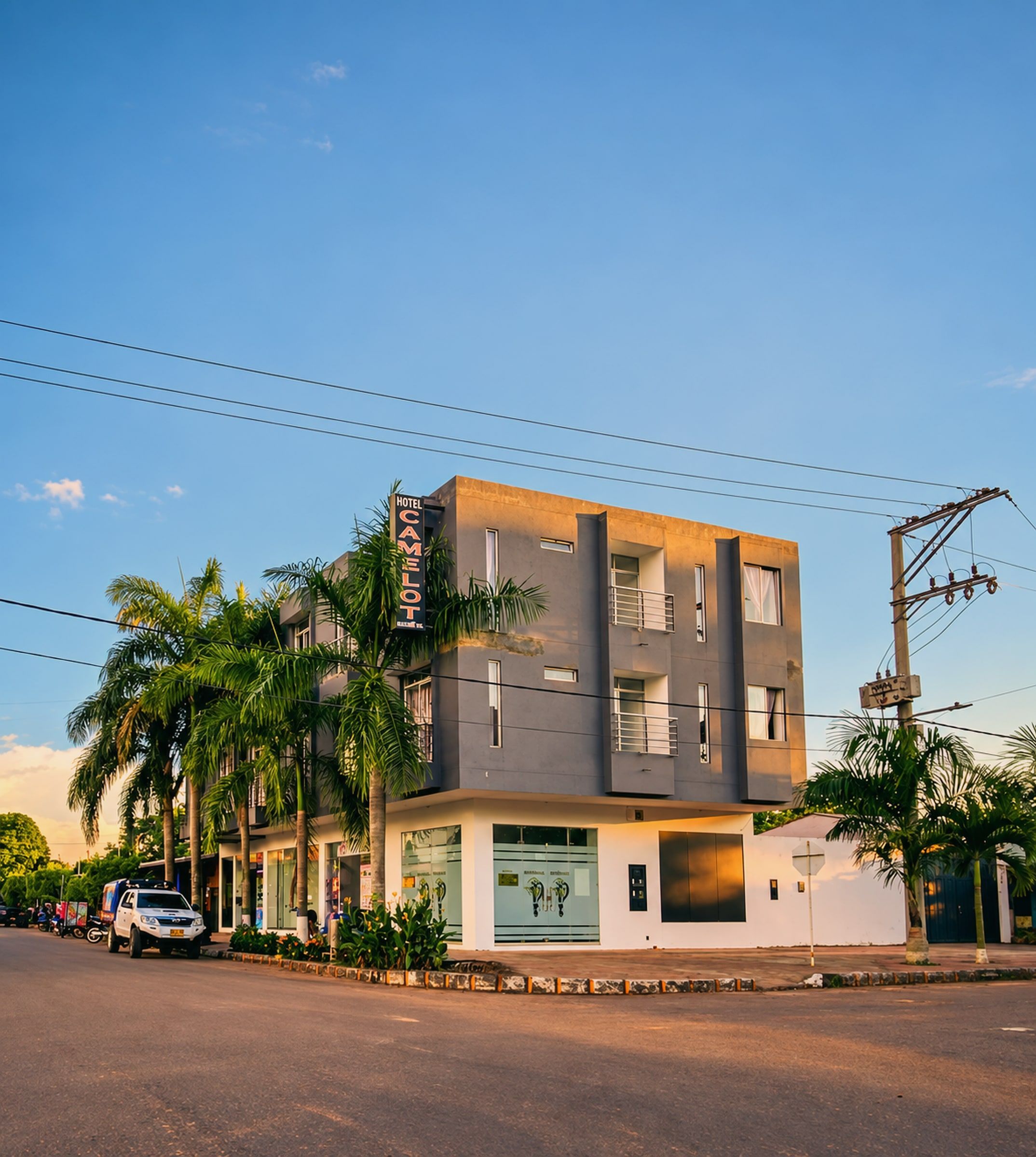 Edificio gris de tres pisos en una esquina, con palmas y el letrero Camelot, iluminado por el sol de la tarde.