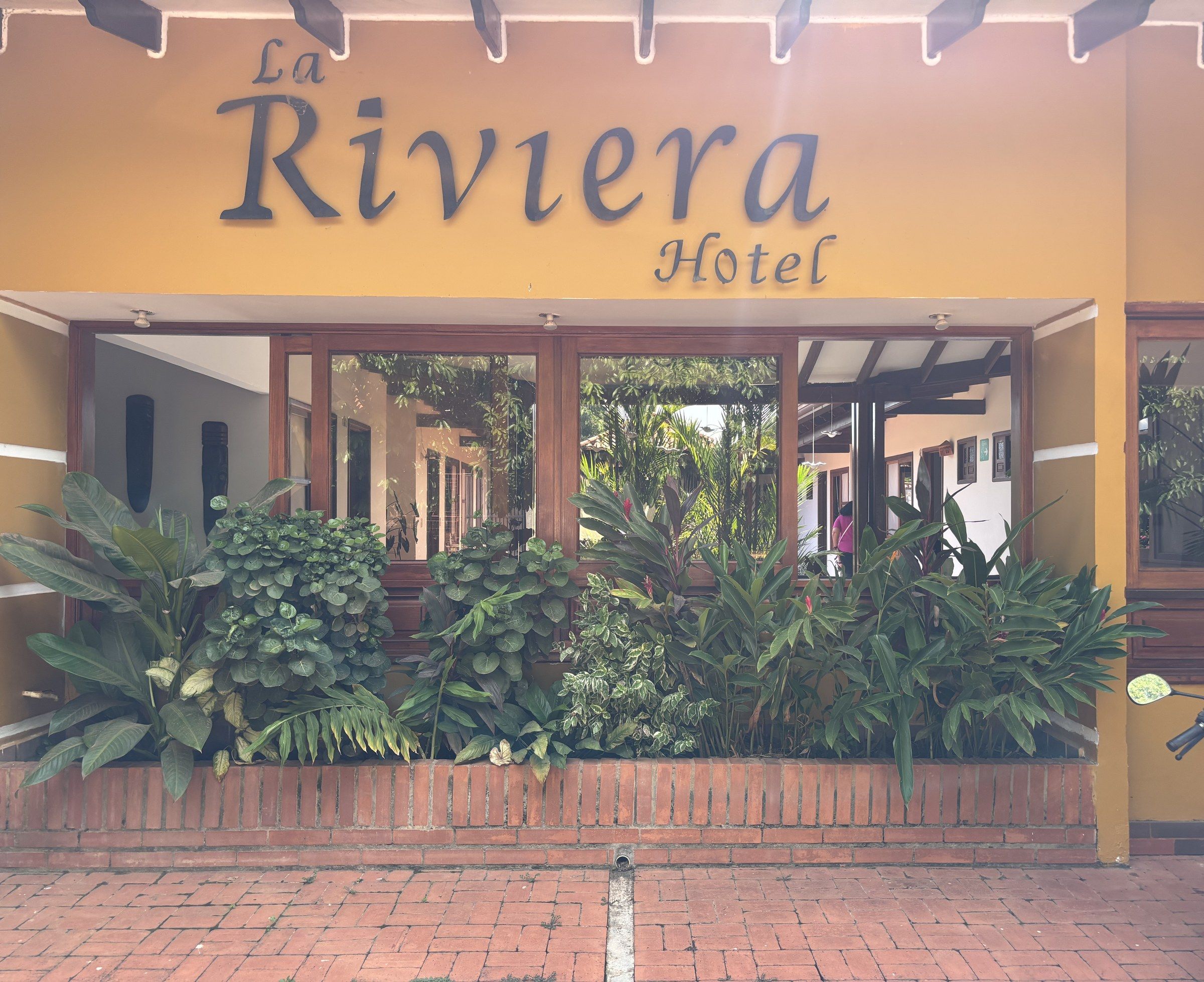 Fachada amarilla de La Riviera con el letrero La Riviera Hotel y un jardín frente a las ventanas.