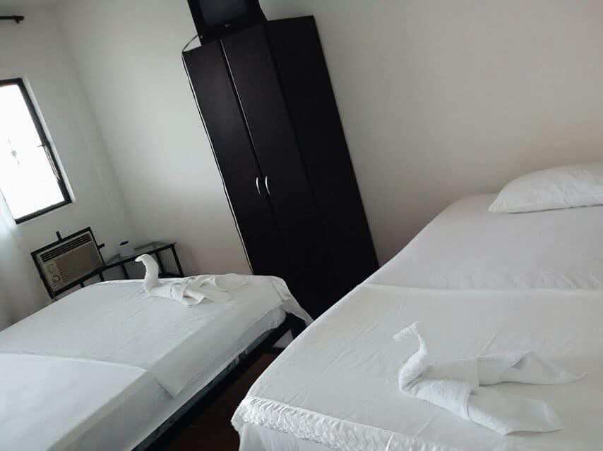 Habitación con dos camas de sábanas blancas y toallas dobladas, aire acondicionado de ventana y un armario negro.