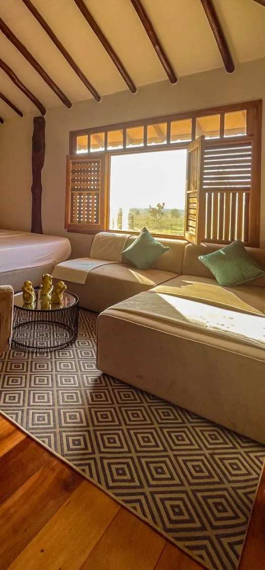 Interior de una cabaña: sofás claros, alfombra y una ventana de madera abierta a la sabana.
