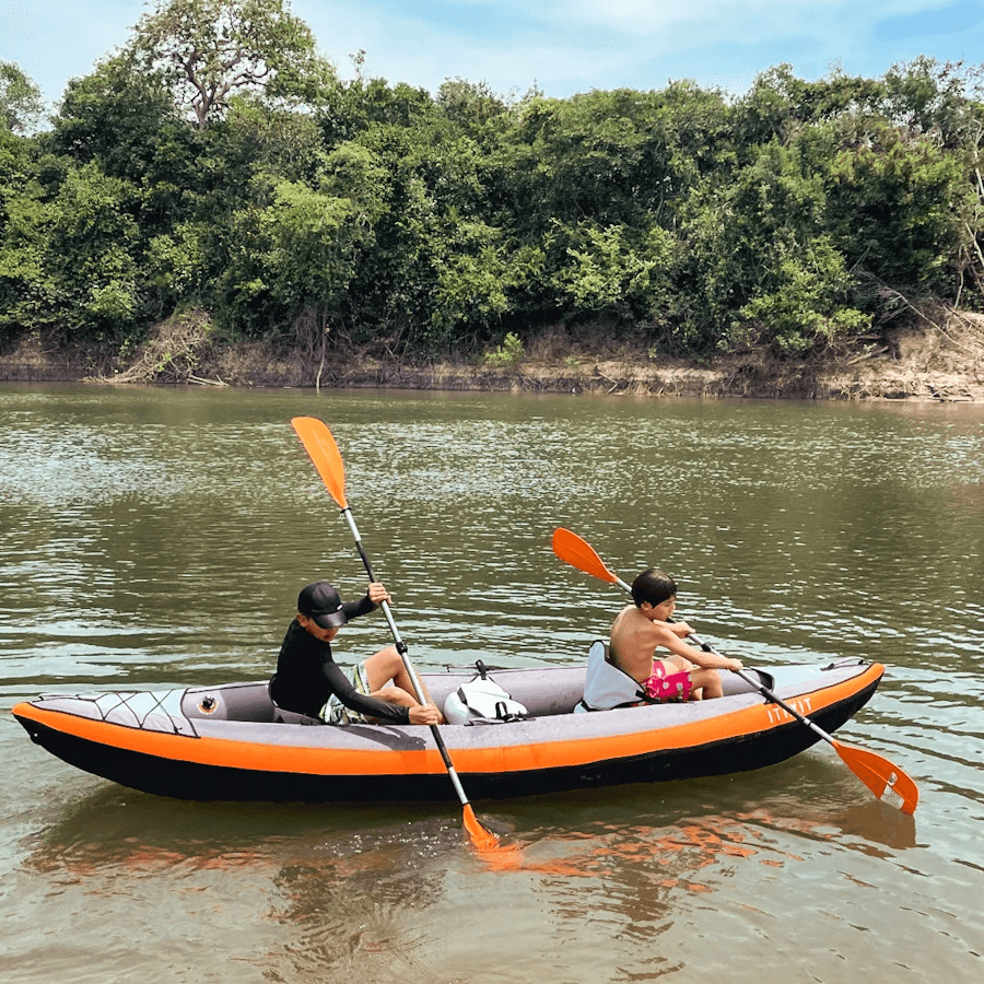 Pareja en un kayak inflable naranja, con los remos en alto, frente a la orilla de monte.