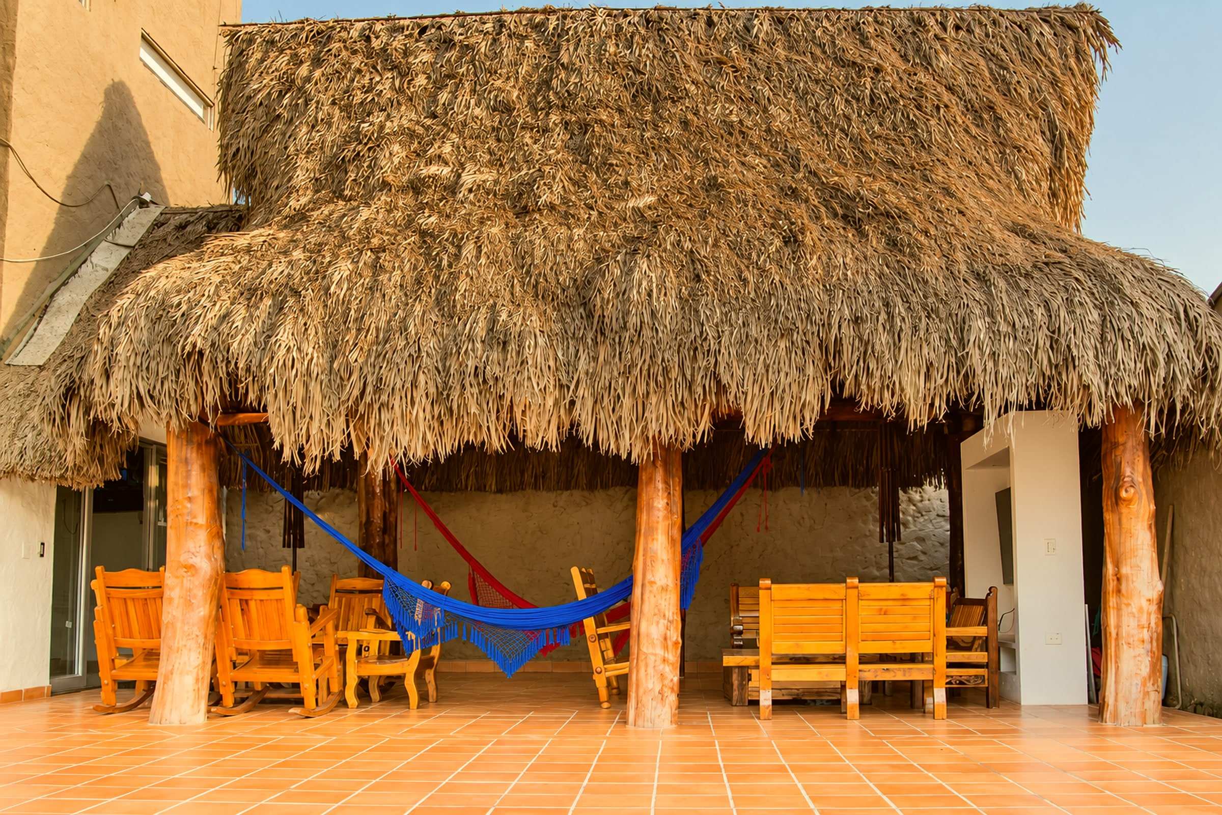 Kiosko con techo de palma, hamaca azul colgada y sillas y bancas de madera.