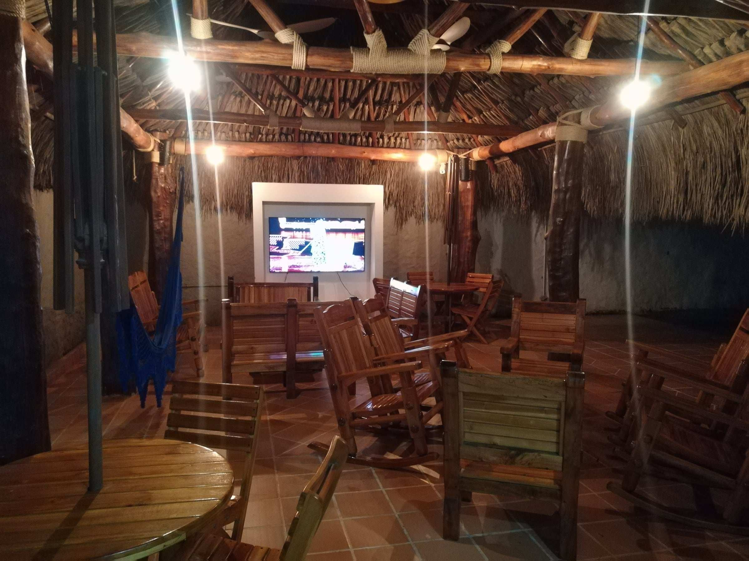 Kiosko con techo de palma iluminado de noche, con televisor, mesas y sillas de madera.