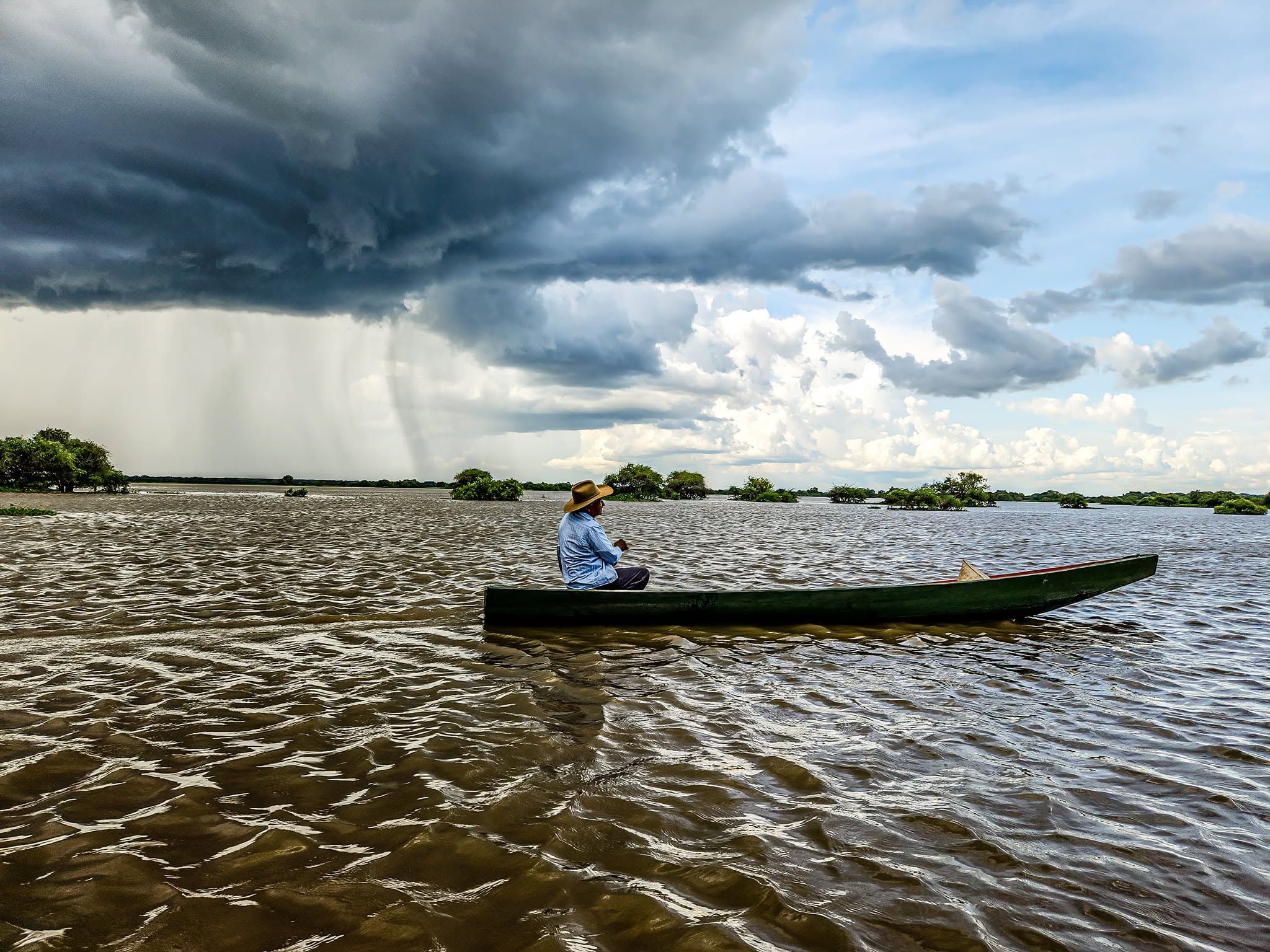 Un hombre con sombrero va sentado en una canoa por la laguna crecida, mientras una tormenta cae sobre la sabana al fondo.