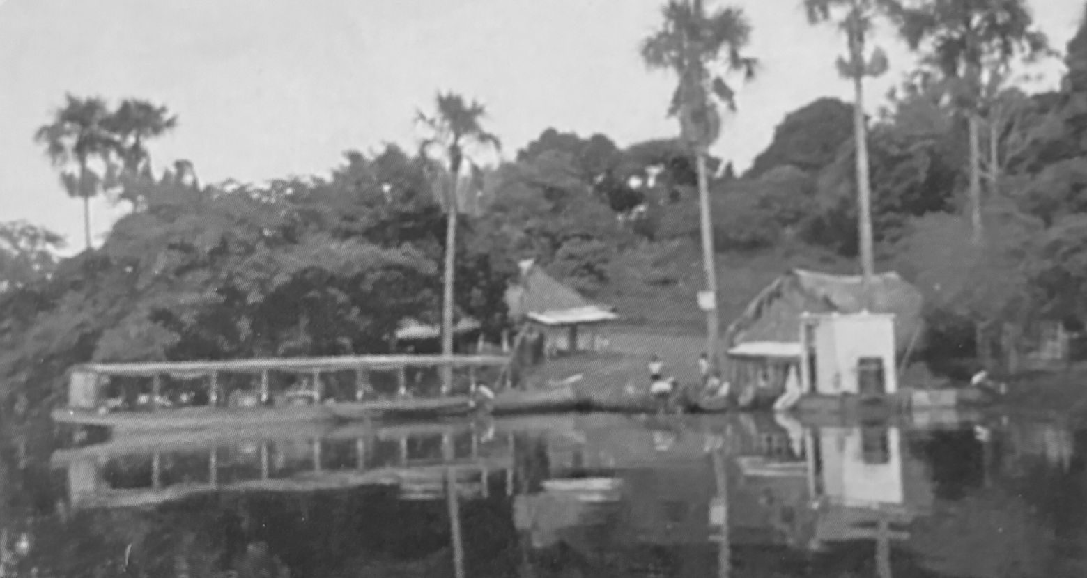 Fotografía antigua en blanco y negro de Laguna La Primavera: un bongo con techo amarrado en la orilla, casas de palma, palmas altas y personas junto al agua.
