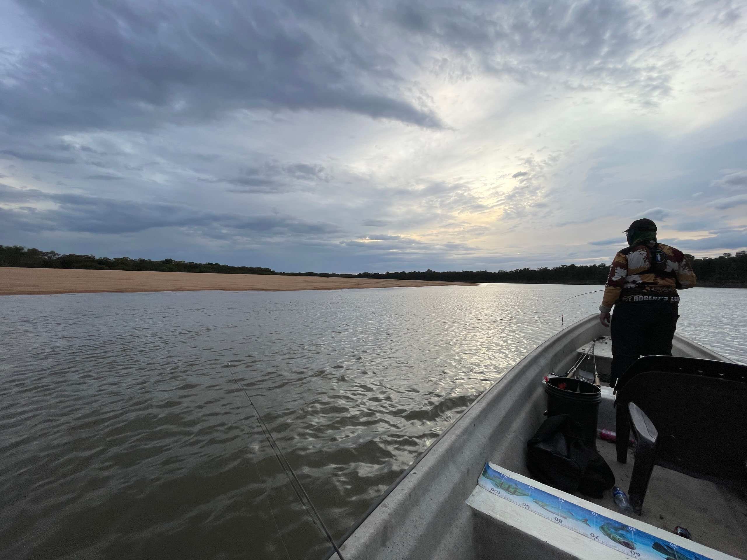 Pescador de pie en la proa de una lancha, lanzando sobre el río bajo un cielo nublado.
