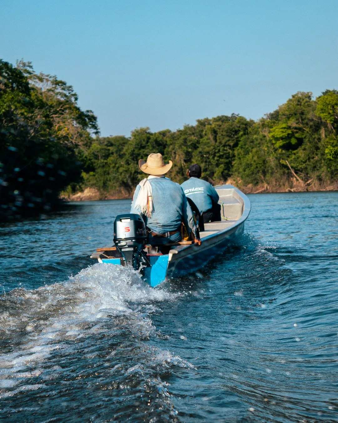 Dos pescadores salen en una lancha con motor por el río, entre el monte de las orillas.