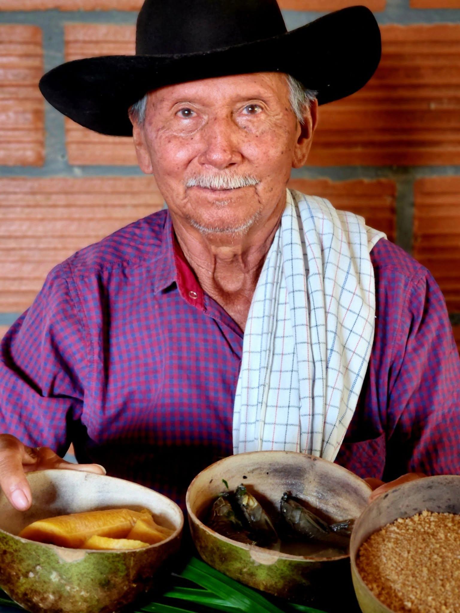 Un llanero mayor, de sombrero negro y camisa de cuadros, ofrece en totumas yuca, caldo de curito y fariña.