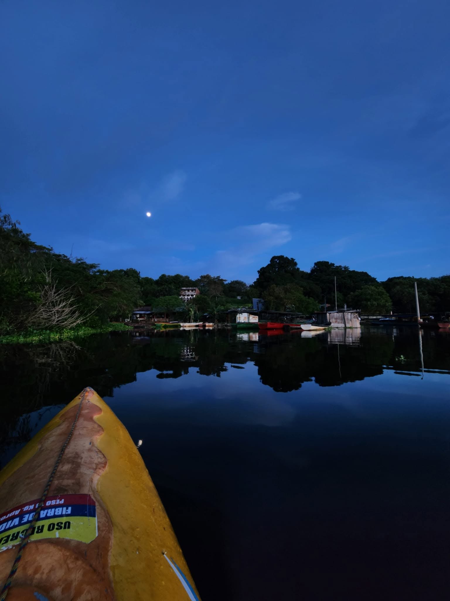 Proa de una canoa en la laguna al anochecer, con el pueblo reflejado.