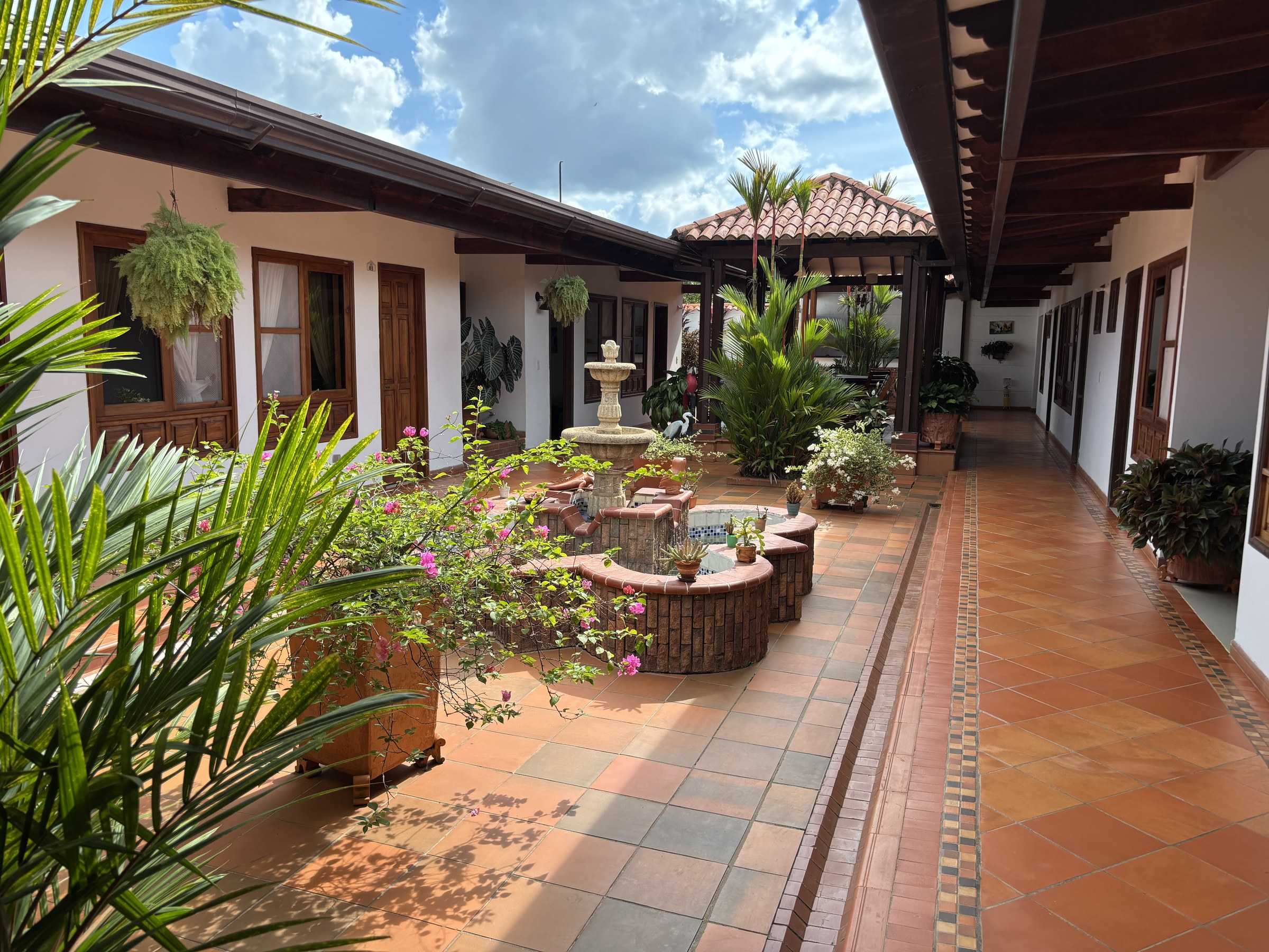 Patio interior con la fuente, palmas y matas en flor, y el corredor de piso de barro bajo el cielo azul.