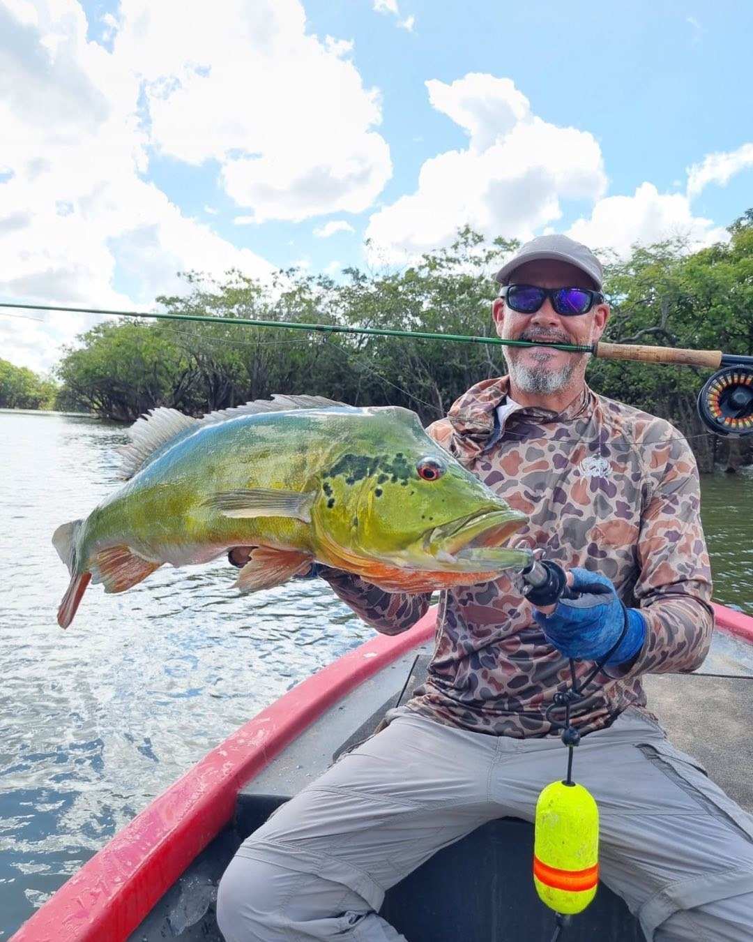 Pescador con gafas oscuras sostiene un pavón verde y amarillo sentado en una lancha roja.