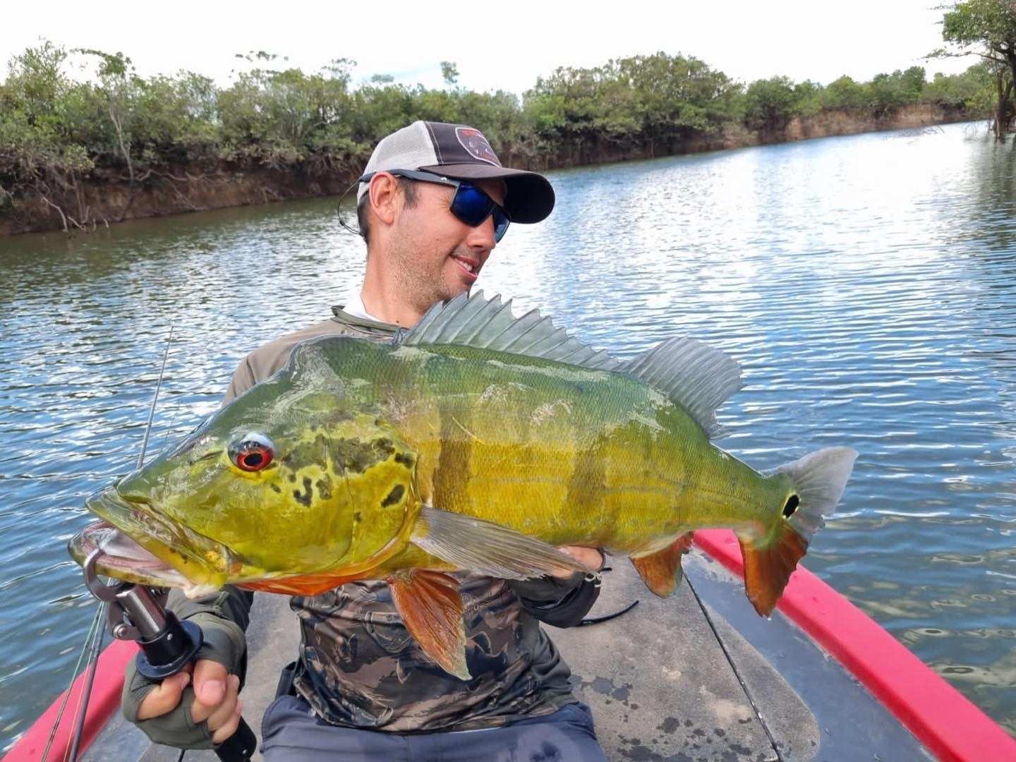 Pescador sentado en una lancha roja sostiene un pavón grande, con el río detrás.