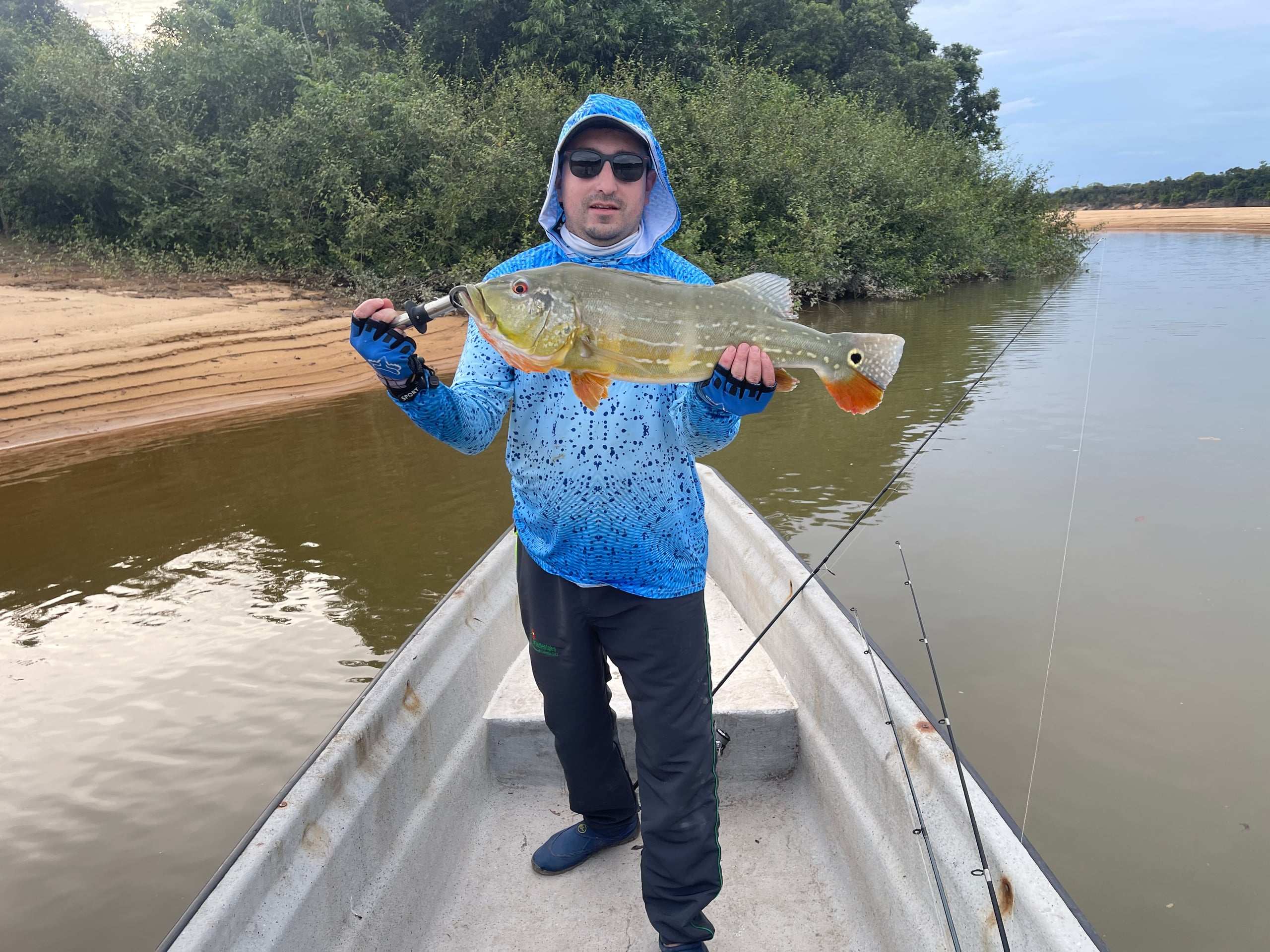 Pescador de pie en la proa de una lancha muestra un pavón, con una playa de arena detrás.