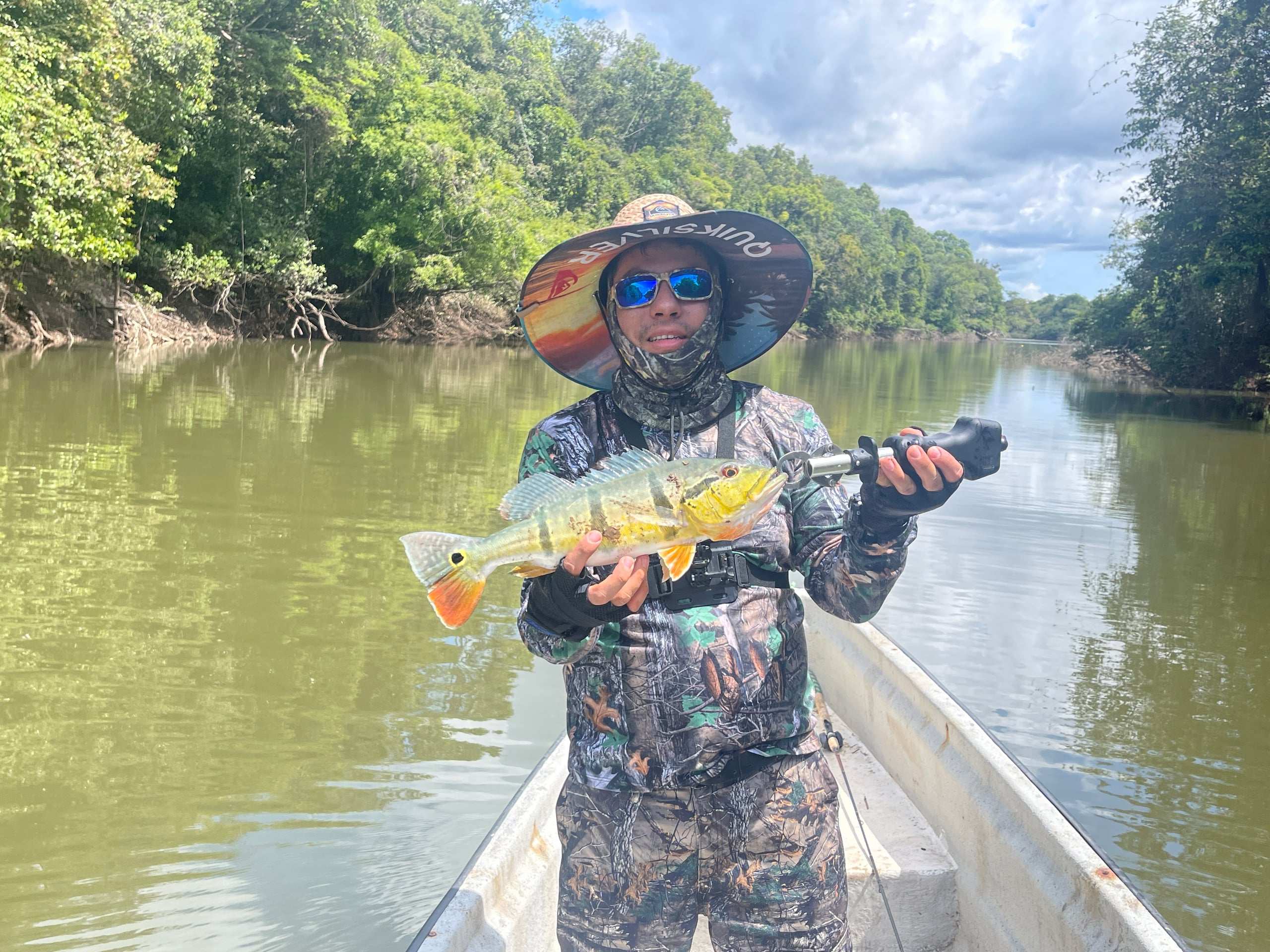 Pescador con sombrero de ala ancha muestra un pavón en la lancha, con el río y el monte detrás.