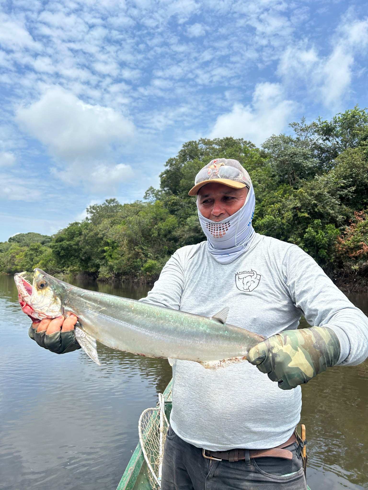 Pescador en una curiara sostiene una payara larga y plateada, con el monte de la orilla detrás.