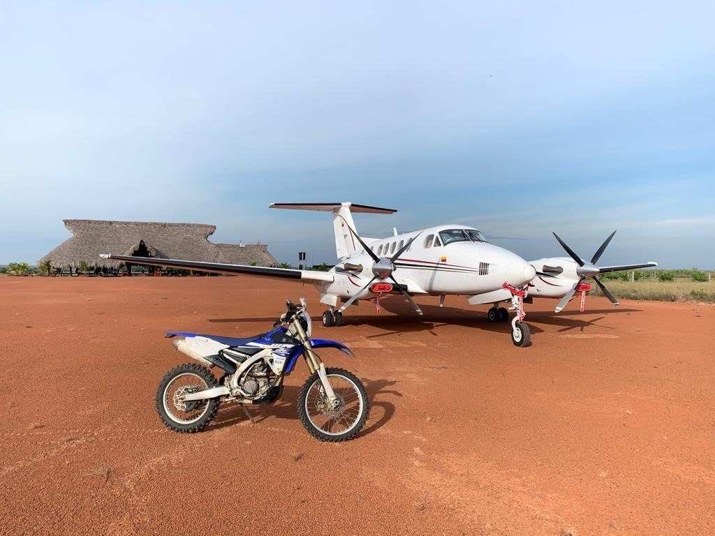 Avioneta blanca estacionada en la pista de tierra roja de Native, con una moto al frente y el kiosco de palma al fondo.