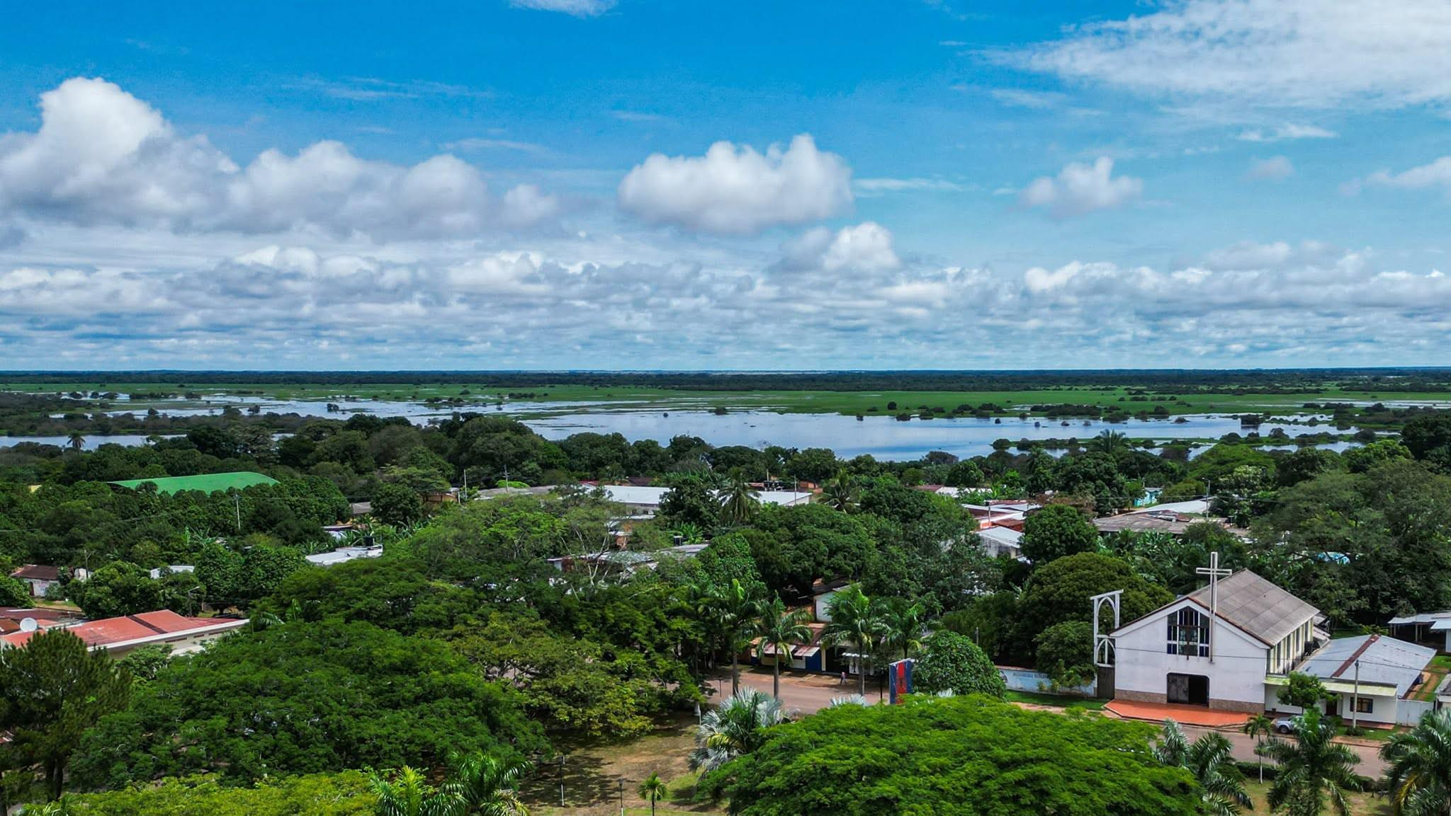 Vista aérea de La Primavera: la iglesia blanca entre árboles y, al fondo, la laguna y la sabana.