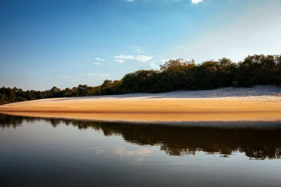 Playa de arena del río al atardecer, con el monte de la orilla reflejado en el agua quieta.