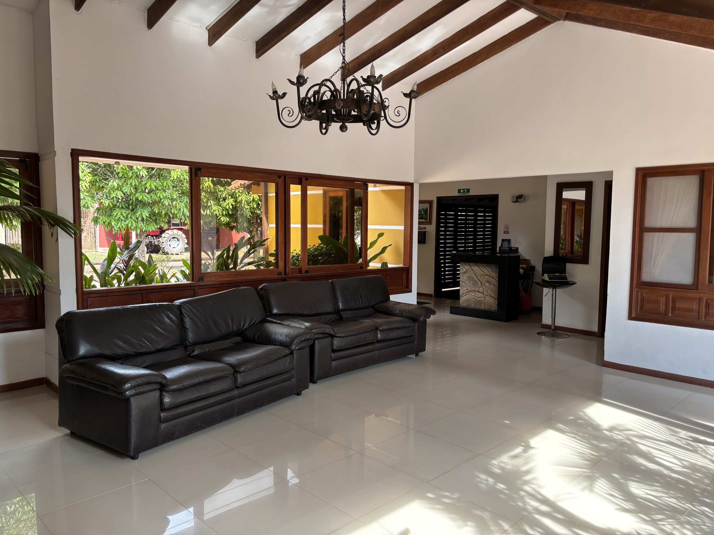 Salón de techo alto con vigas, sofás negros de cuero, lámpara de hierro y ventanales al jardín.